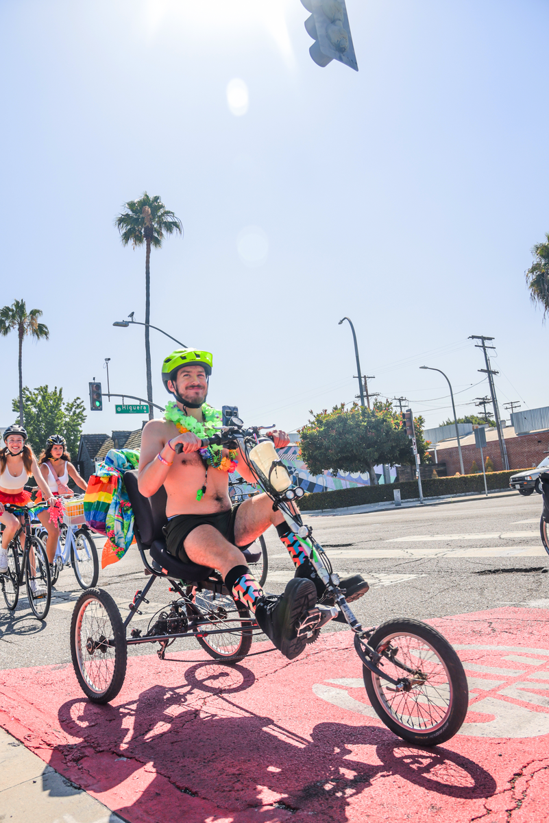 A man wearing rainbow leis, green shorts nad colorful socks is riding a 3-wheel bicycle.