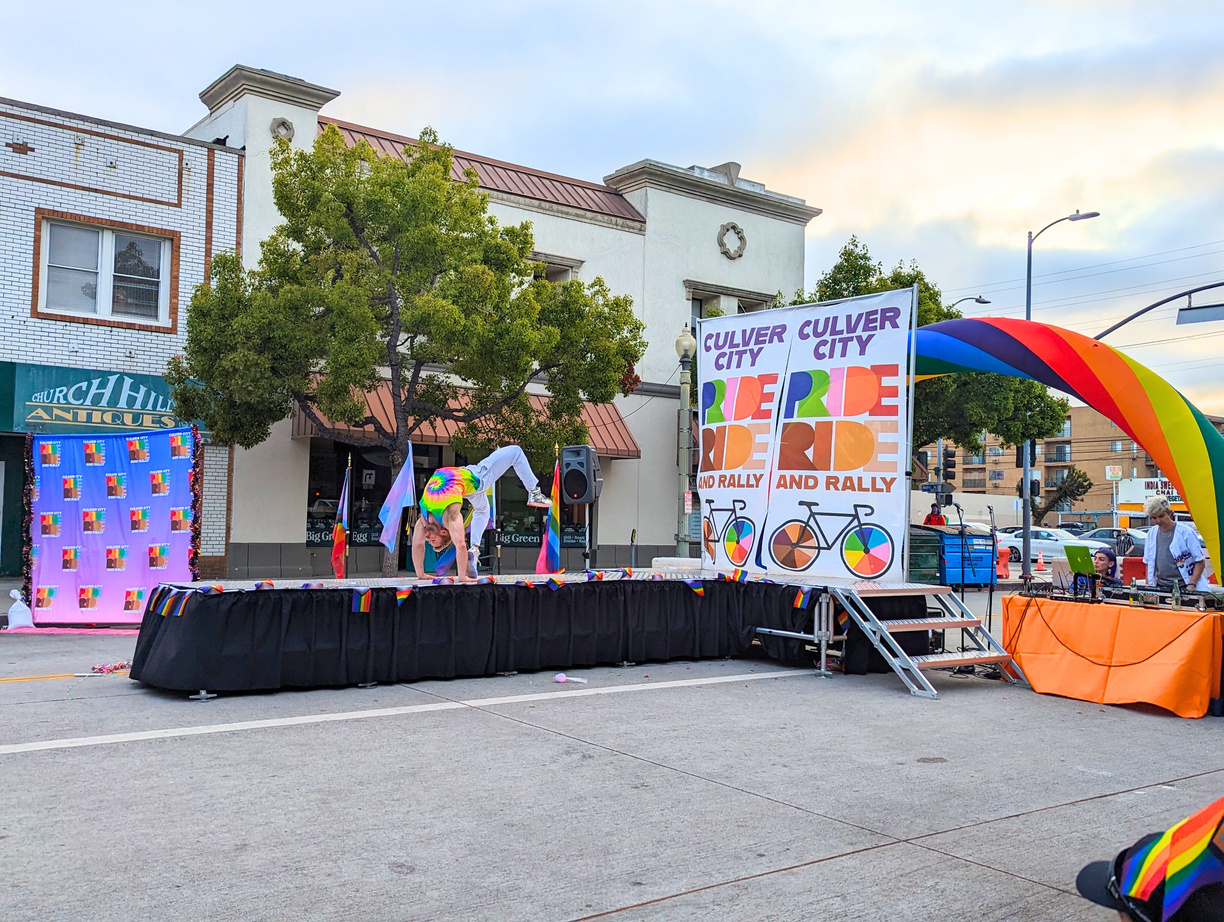 A dancer is mid-air during a performance on the CUlver City Pride platform stage. He is located on the runway portion of the stage. There is a banner at the rear of the stage which states, "CULVER CITY PRIDE RIDE AND RALLY"