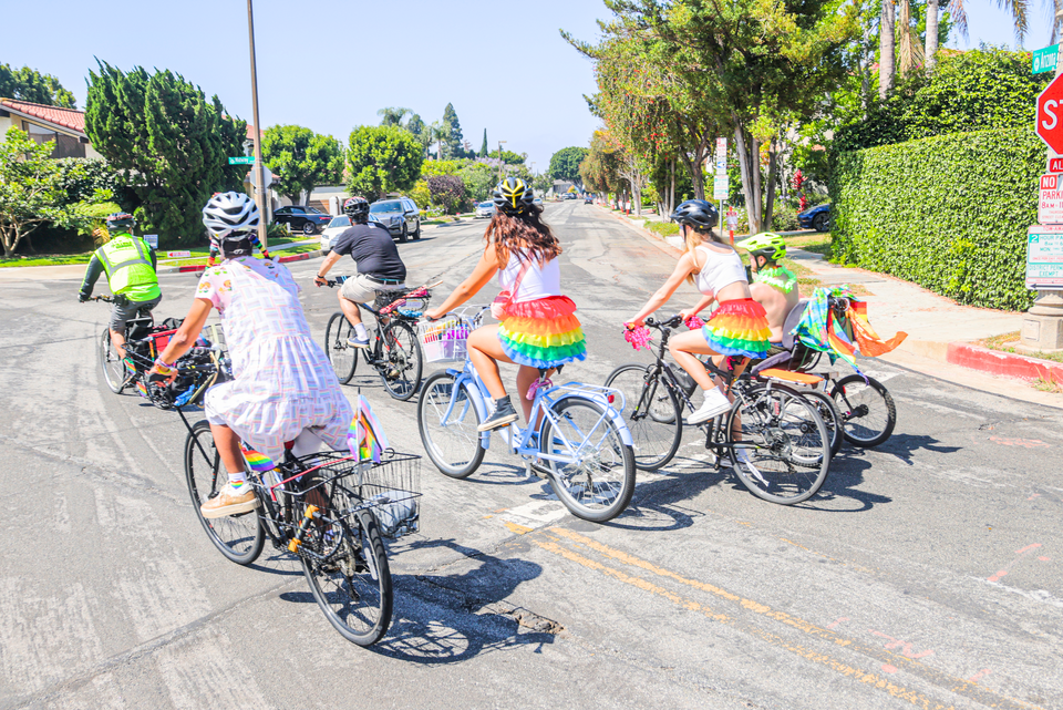 a group of people riding bikes on a residential street wearing rainbow tutus. there are trees in the background.